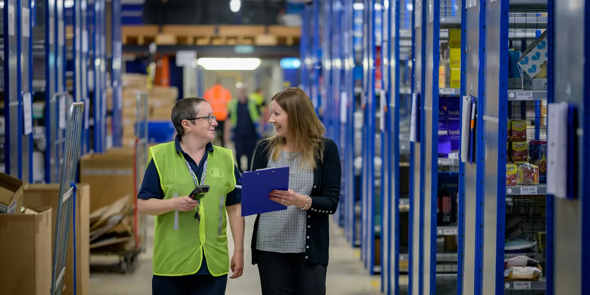 Two colleagues with a clipboard and scanner walk down a warehouse aisle of stocked shelving