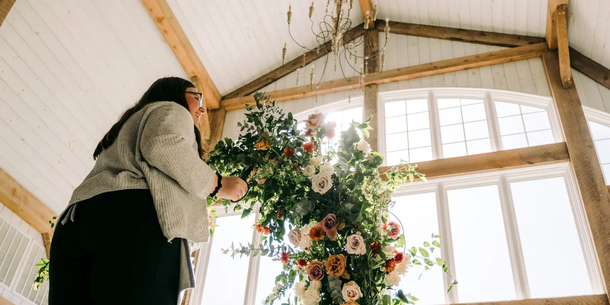 A woman arranges a tall floral installation in a bright, timber-beamed event hall