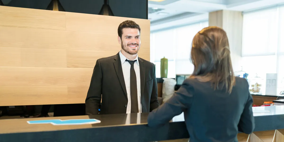 A receptionist welcomes a guest with a suitcase at a hotel front desk