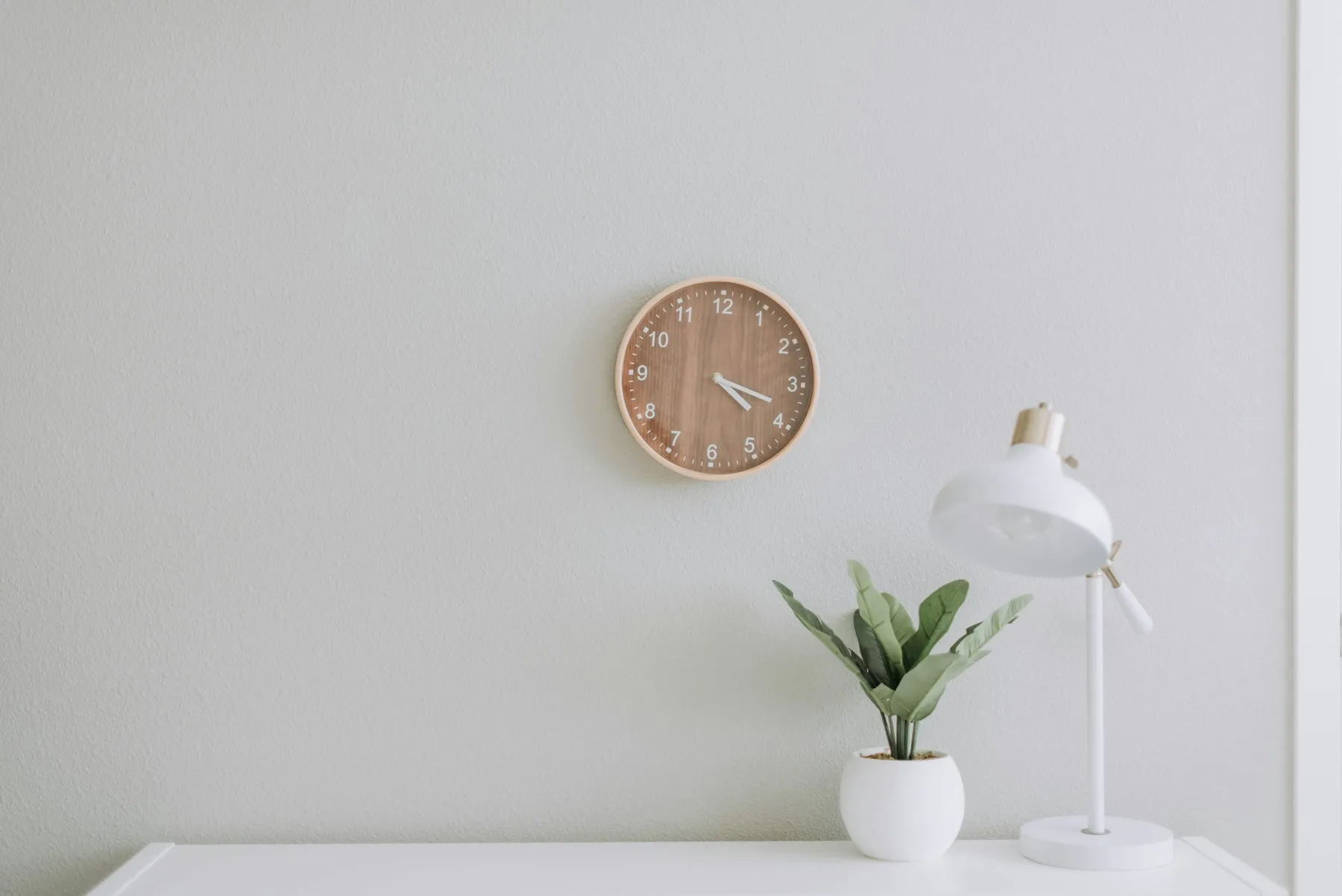 A quiet desk with a clock, a lamp and a plant against a white wall