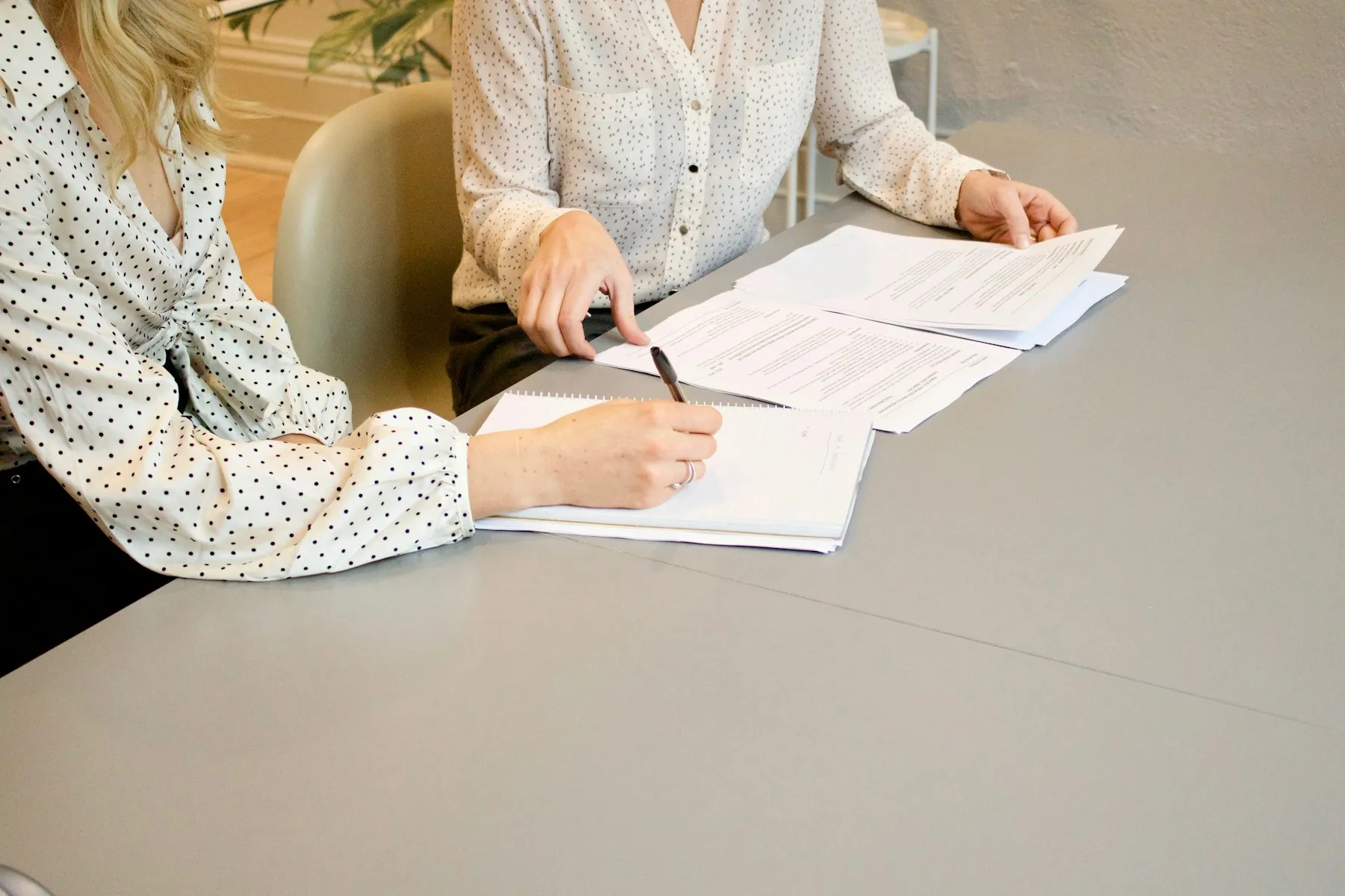 One person signs a document while a colleague reviews the pages beside her