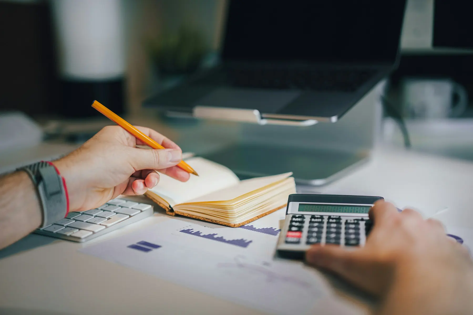 Hands with a pencil and a calculator over a notebook and a printed report, a laptop behind